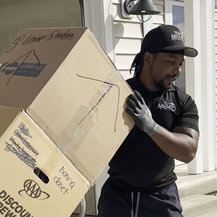 A person wearing a cap and gloves carrying a large cardboard box outside a building.